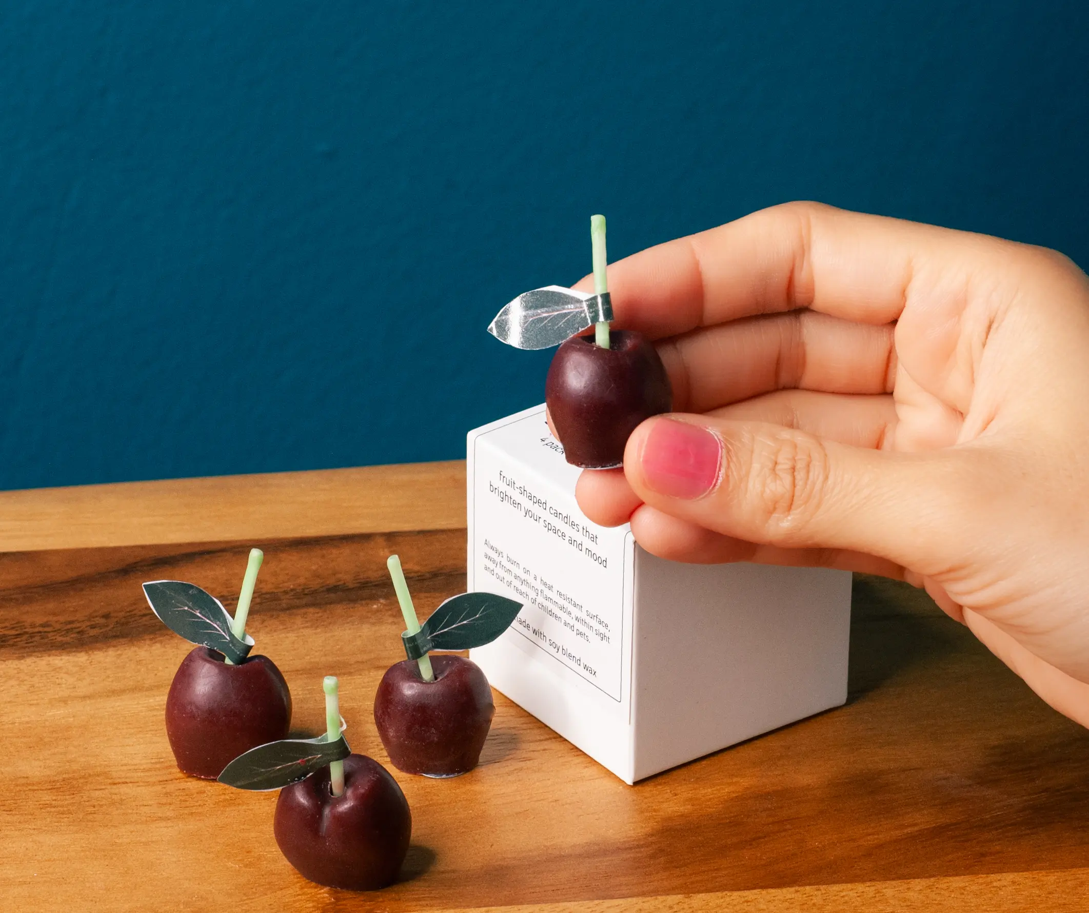 Cute maroon cherry candles kept on the box of candles. It is very small set of candles. A woman holding one cherry candle that is a very realistic candle