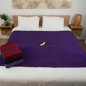 Dark Purple waterproof blanket on a bed and a banana on top of it for scale. There are three folded sex blankets kept on top of the bed showcasing all the colours Banana Passion offers. This banana passion sex blanket is better than splash blanket for intimacy as it is absorbent.