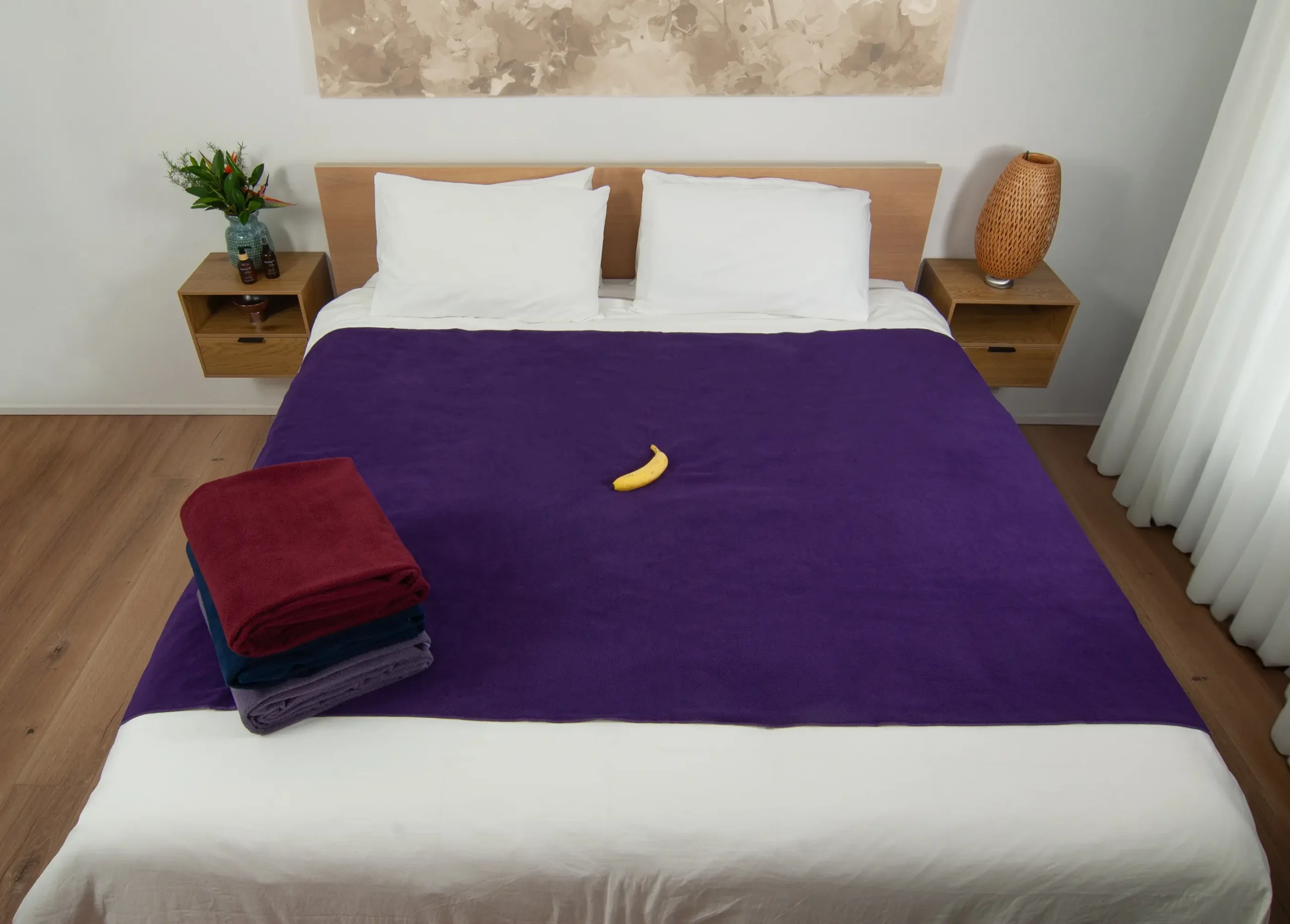 Dark Purple waterproof blanket on a bed and a banana on top of it for scale. There are three folded sex blankets kept on top of the bed showcasing all the colours Banana Passion offers. This banana passion sex blanket is better than splash blanket for intimacy as it is absorbent.