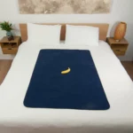 Navy Blue Waterproof Sex blanket on the bed with a banana used for scaling in a bedroom. Two bed side tables in this room. One table has massage oils and a vase with Australian natives in it. One side table has an ikea lamp on it.