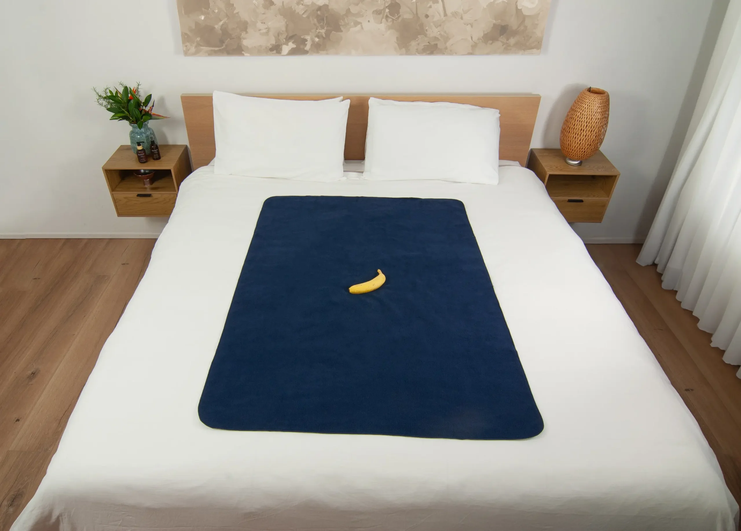 Navy Blue Waterproof Sex blanket on the bed with a banana used for scaling in a bedroom. Two bed side tables in this room. One table has massage oils and a vase with Australian natives in it. One side table has an ikea lamp on it.