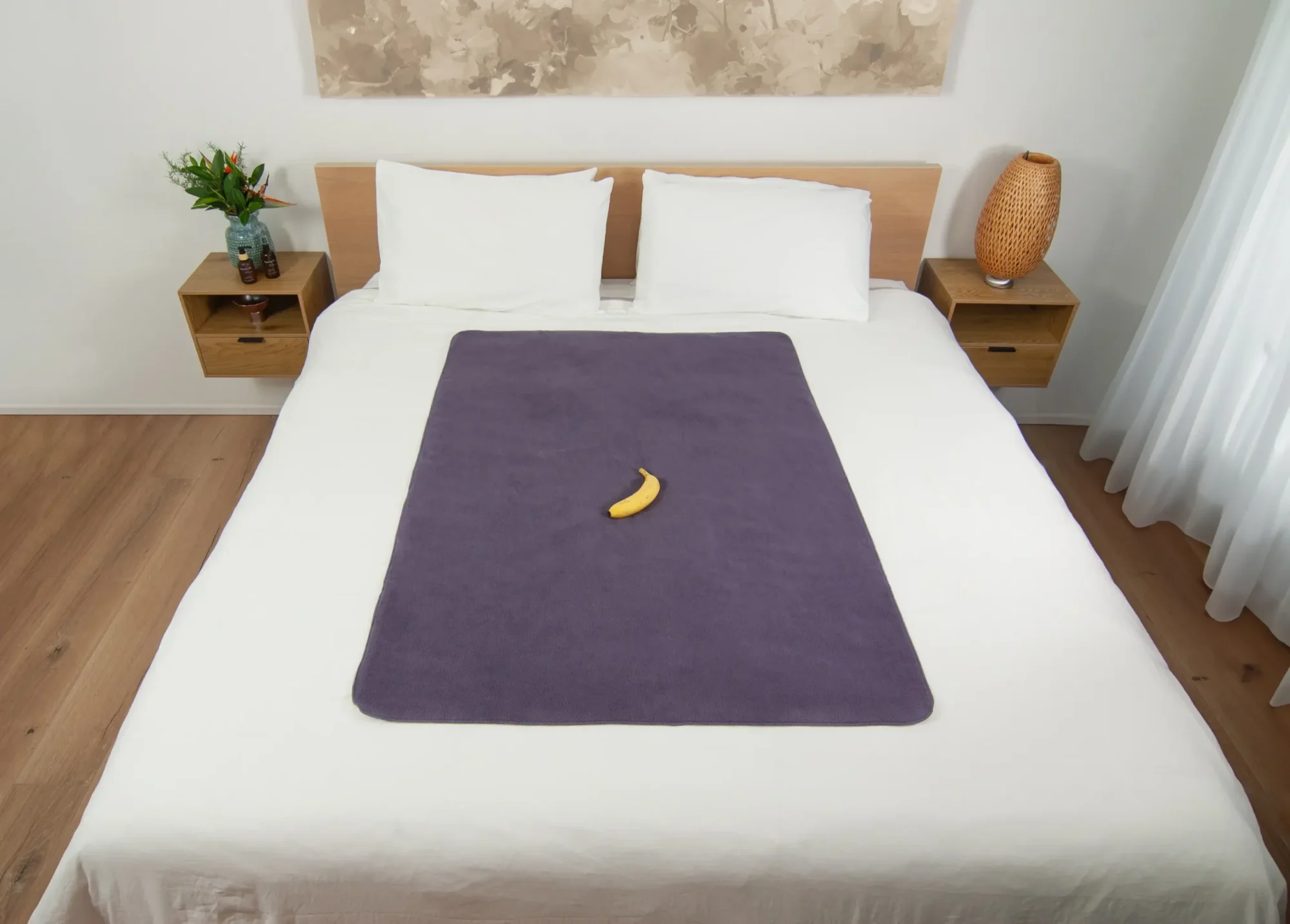 Heather Grey Waterproof Sex blanket on the bed with a banana on top it for scale. There is a painting on the wall and there are two floating bed side tables.