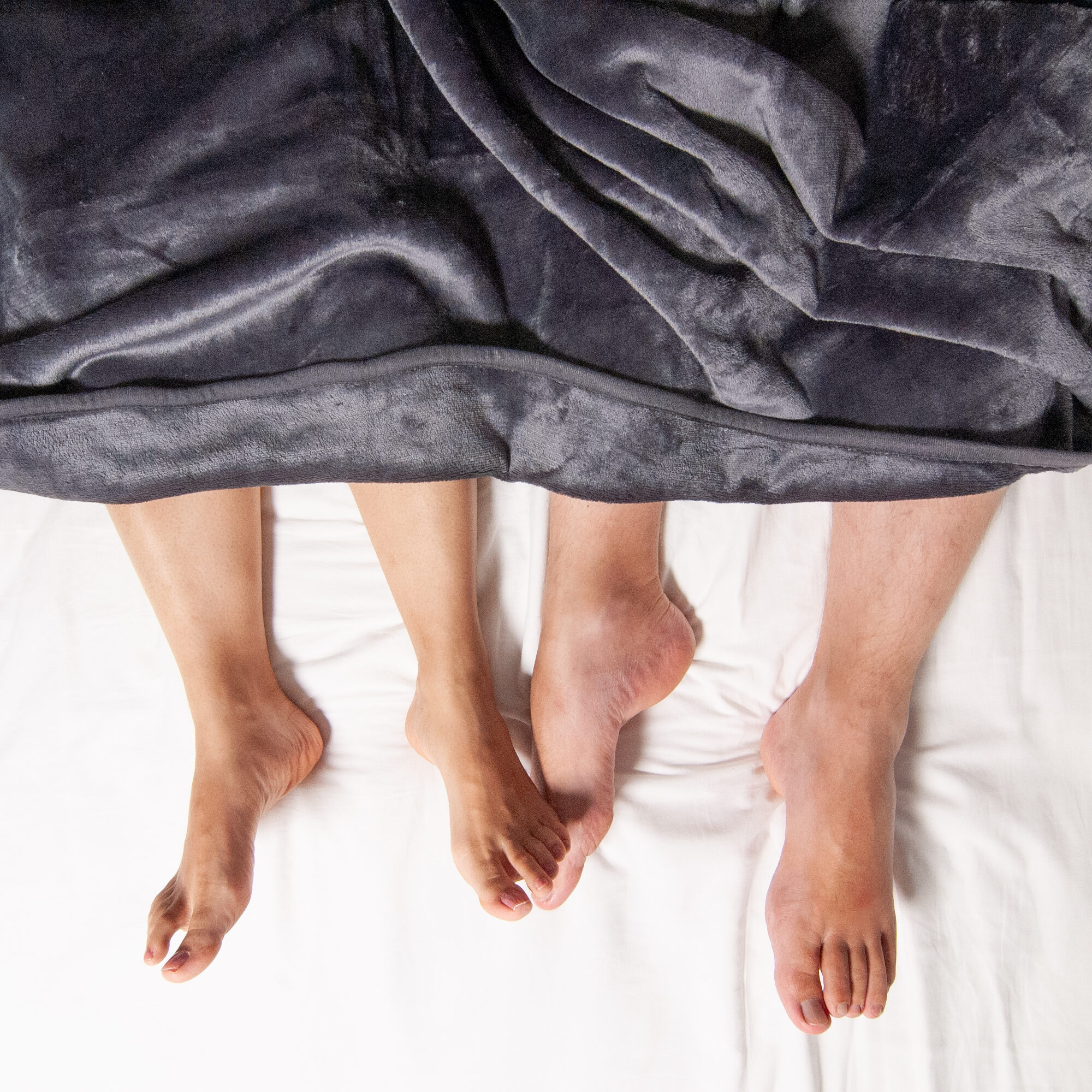 feet of two people shown underneath a Banana Passion bright red velvety waterproof blanket, with bedsheets shown underneath