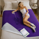 A young woman lies down on a bed, on top of a purple GripMe waterproof blanket while she pours water from a glass. The water splashes and gets absorbed into the blanket. A large tag attached to the blanket mentions anti-slip to describe the silicon dotted backside.