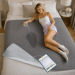 A young woman lies down on a bed, on top of a grey GripMe waterproof blanket while she pours water from a glass. The water splashes and gets absorbed into the blanket. A large tag attached to the blanket mentions anti-slip to describe the silicon dotted backside.